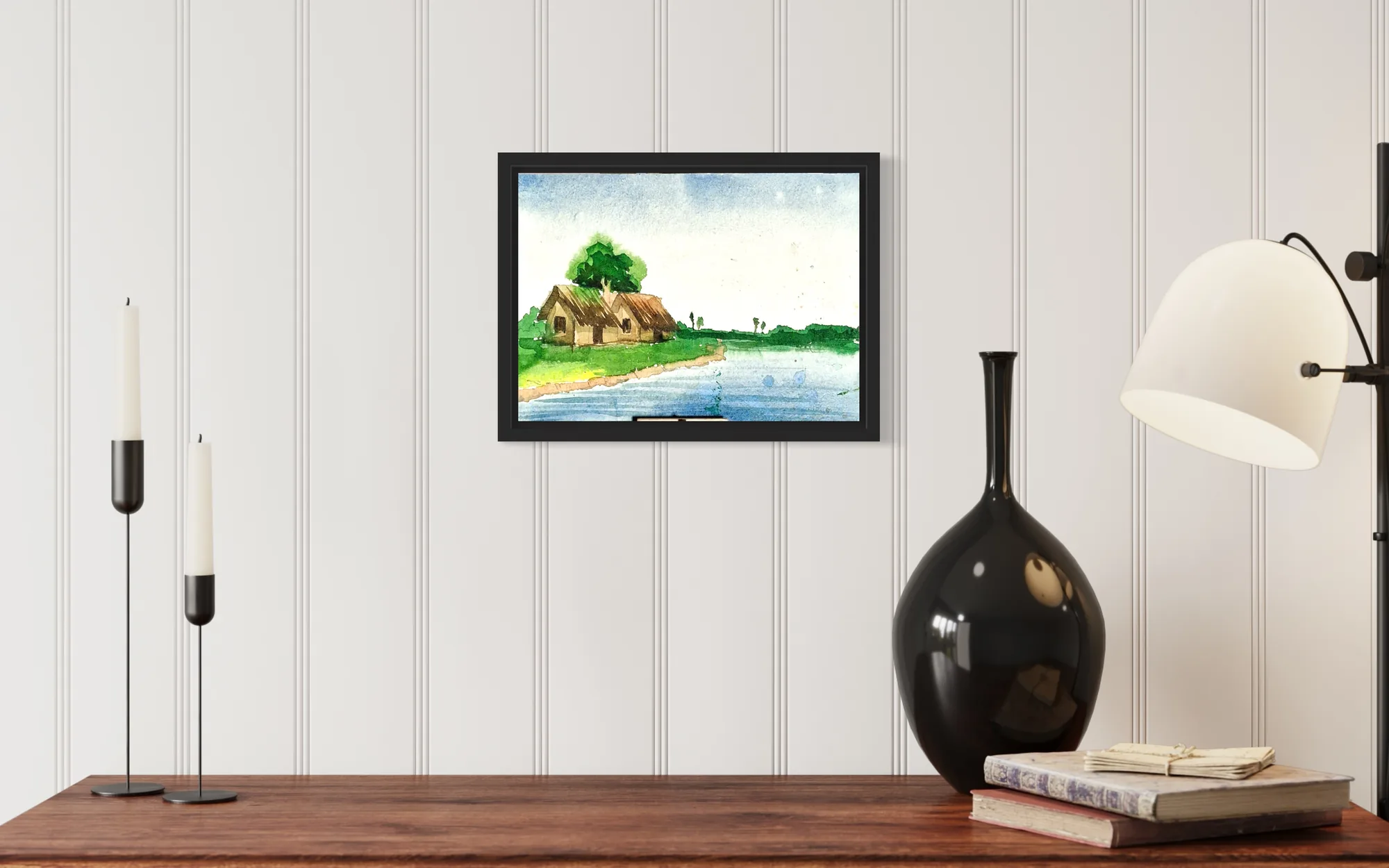Watercolor scene of green huts by a blue river under a bright sky, in a black frame.